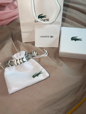 Silverfärgat armband från Lacoste - Stilrent armband i silverfärgad metall från Lacoste med länkdesign och ingraverad krokodil-logga på flera länkar. Kommer med originalpåse, ask och påse från Lacoste – perfekt för dig som gillar snygga accessoarer med sportig touch.
