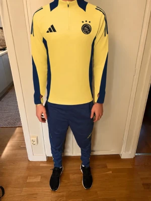 Ajax träningsoverall gul/blå Adidas - Ajax träningsoverall från Adidas i gult och blått. Överdel med halv dragkedja, klubbmärke och Adidas-logga på bröstet samt tre stjärnor. Matchande blå byxor med klubbmärke och gula detaljer. Perfekt för fotbollsträning eller chill. Personen i bilden är 184 cm, liten fläck med som man ser på bilden 