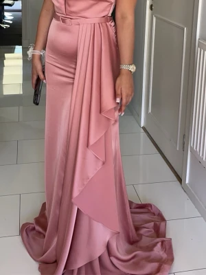 Lång rosa offshoulder klänning satin - Elegant långklänning i rosa satin med offshoulder-ärm och figurnära passform. Klänningen har ett vackert fall med draperad volangdetalj framtill och ett bälte i midjan. Perfekt för speciella tillfällen. Perfekt för som bröllopsgäst, för bal och speciella tillfällen, klänningen passar en 36 också🥰