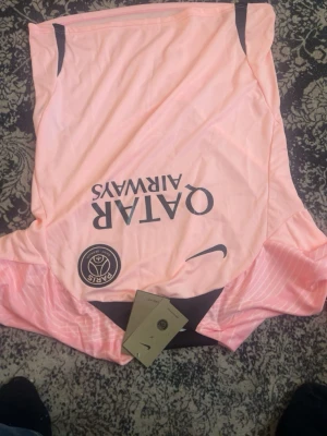 PSG rosa matchtröja från Nike - Säljer en rosa Paris Saint-Germain matchtröja från Nike med svarta detaljer och Qatar Airways-tryck på bröstet. Tröjan har korta ärmar och PSG-logga på vänster sida. Perfekt för fotbollsträning eller att bära som supporter.