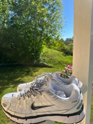Nike P-6000 vita sneakers - Nike P-6000 sneakers i vitt med svarta detaljer och silvriga inslag. Skorna har en sportig mesh- och syntetöverdel, svart swoosh-logga på sidan och P-6000-tryck. Sulan är vit med svarta partier och snörningen är vit med svarta ränder.