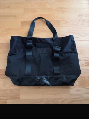 Svart shoppingväska från Gymshark - Stor svart totebag från Gymshark med breda axelband och detaljer i glansigt material längst ner. Väskan har dragkedja upptill och rymligt huvudfack, perfekt för gymmet eller vardagsgrejer. Passar att ha över en helg eller under sätet på flyget när man reser! Den är 50x35x15