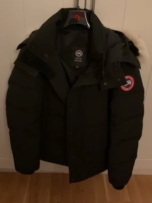 Svart Canada Goose Wyndham - Säljer en svart parkas från Canada Goose med klassisk patch på ärmen och stor huva med äkta päls. Jackan är dunfodrad och har en rak, puffig passform som håller dig varm under kalla dagar. Perfekt för vintern och riktigt snygg streetstil.