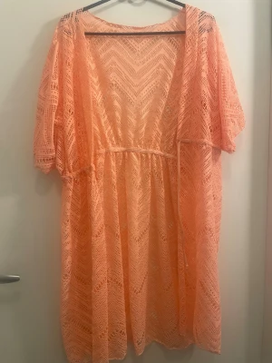 Persikofärgad strandkimono i spets - Snygg persikofärgad strandkimono i genombruten spets med knytband i midjan. Lätt och luftig modell med korta ärmar och vackert mönster i tyget. Perfekt att slänga över bikinin på stranden eller vid poolen.