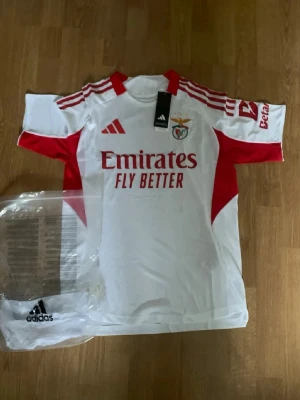 Benfica matchtröja - Snygg Benfica matchtröja från Adidas i vitt med röda detaljer och klubbmärke på bröstet. Tröjan har röda ränder på axlarna, röd krage och sponsortryck framtill. Tillverkad i lätt och ventilerande material, perfekt för fotbollsträning eller match.