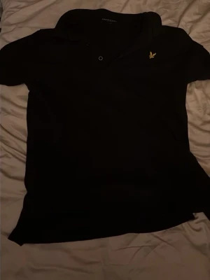 Svart pikétröja från Lyle & Scott - Klassisk svart pikétröja från Lyle & Scott med gul logga på bröstet. Tröjan har krage, korta ärmar och knäppning framtill. Perfekt för en stilren och avslappnad look.
