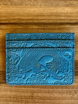 Blå korthållare från De’Jour - Snyggt blått kortfodral från Dé Jour i slätt och präglat läder. Framsidan har ett detaljerat fiskmotiv och blommönster, medan baksidan är stilren med logotyp. Perfekt för kort och ID, smidig att ha i fickan.