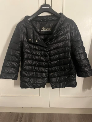 Svart dunjacka från Herno - Snygg svart dunjacka från Herno med glansig finish och horisontella sömmar. Jackan har trekvartsärmar, tryckknappar och en stilren passform. Tunn och perfekt nu inför sommar/ vår! Den har en liten defekt på ryggen men inget som syns tydligt.