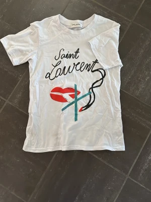 Vit Saint Laurent t-shirt med tryck - Säljer en vit t-shirt från Saint Laurent med coolt grafiskt tryck framtill. Klassisk passform och rund halsringning. Perfekt för dig som vill ha en statement tee från ett ikoniskt märke. Tröjan är liten i passformen.