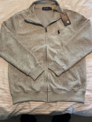 Grå zip hoodie från Polo Ralph Lauren - Snygg grå zip hoodie från Polo Ralph Lauren med klassisk svart logga broderad på bröstet. Tröjan har dragkedja hela vägen, ribbade muddar och två fickor framtill. Tillverkad i mjukt bomullsmaterial för en skön känsla.
