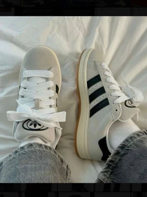 Adidas Campus 00S W - Snygga Adidas Campus sneakers i grå mocka med svarta klassiska ränder på sidorna och vit sula med gummidetalj. Tjocka vita skosnören (ingår även svarta tjocka skosnöre) och stor Adidas-logga på plösen. Köpta för 1449kr.