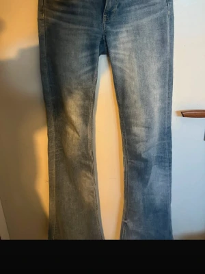 Ljusblå bootcut jeans - Säljer ett par ljusblå bootcut jeans med slitna detaljer och klassiska bakfickor. Jeansen har en lätt utsvängd passform nertill och är tillverkade i denim. Perfekta för dig som gillar en avslappnad och trendig stil.