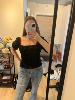 Svart offshoulder blus med volang - Trendig svart blus med offshoulder och korta puffärmar. Toppen har smockad midja och volangdetalj längs halsringningen. Perfekt att styla med jeans för en cool look.