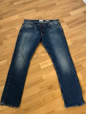 Blå raka jeans från Replay - Säljer ett par klassiska blå jeans från Replay med raka ben och snygga slitningar framtill. Jeansen har fem fickor, kontrastsömmar och en cool tvättad look. Perfekta för dig som gillar en avslappnad stil.