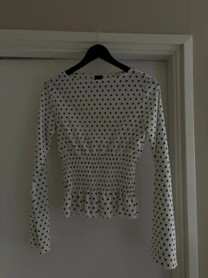 Prickig långärmad topp Gina Tricot - Superfin vit långärmad topp från Gina Tricot med svarta prickar. Toppen har smockad midja som ger en snygg form och lätt volang nertill. Perfekt att styla med jeans eller kjol för en trendig look.
