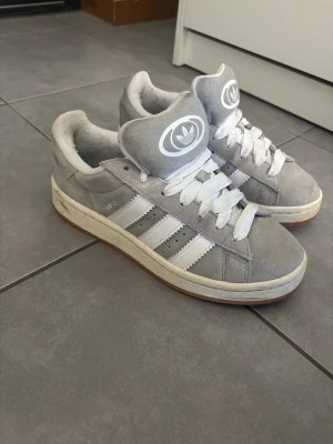 Grå Adidas Campus sneakers - Snygga grå Adidas Campus sneakers med klassiska vita ränder på sidorna och vit logga på plösen. Ovandelen är i mocka och sulan är vit med en gummidetalj. Perfekta för en streetwear-look och passar till det mesta.