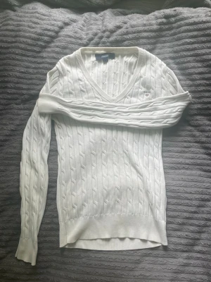 Vit tröja från GANT - Vit kabelstickad tröja från GANT med V-ringning och broderad logga på bröstet. Tröjan har långa ärmar, ribbade muddar och klassisk passform. Perfekt för en stilren och tidlös look.