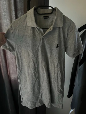 Grå pikétröja från Polo Ralph Lauren - Klassisk grå pikétröja från Polo Ralph Lauren med korta ärmar och krage. Tröjan har den ikoniska broderade loggan på bröstet och är gjord i mjuk bomull för en bekväm känsla. Perfekt till jeans eller shorts för en clean look. I princip helt ny