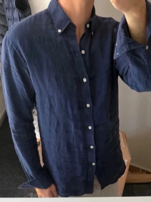 Mörkblå linneskjorta från John Henric  - Snygg mörkblå skjorta i 100% linne från John Henric med klassisk krage och vita knappar. Skjortan har lång ärm och en avslappnad passform, perfekt för en stilren look. Enkel och tidlös design som funkar till många outfits.