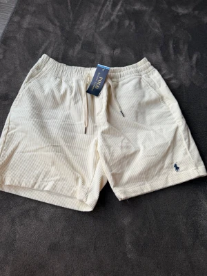 Vita cordshorts från Polo Ralph Lauren - Snygga vita shorts i tunn manchester från Polo Ralph Lauren. De har resår i midjan med dragsko och två sidofickor. Klassisk blå logga broderad på ena benet. Perfekta för en avslappnad och stilren look.