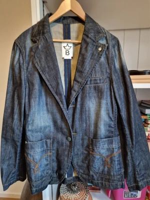 Jeansblazer från Bläck med fickor - Snygg jeansblazer från Bläck i mörkblå denim med klassisk krage och två stora framfickor med dekorativa sömmar. Kavajen har en bröstficka och stängs med en knapp framtill. Perfekt för en avslappnad men stilren look.