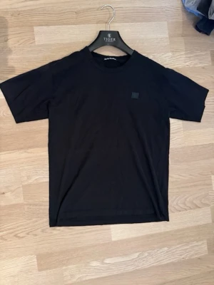 Svart t-shirt från Acne Studios - En stilren svart t-shirt från Acne Studios med klassisk rund hals och diskret logopatch på bröstet. T-shirten har korta ärmar och är gjord i mjuk bomull.