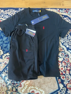 Svart set från Polo Ralph Lauren - Säljer ett svart set från Polo Ralph Lauren med t-shirt och shorts. Båda plaggen har det klassiska röda Polo-logot på bröstet och benet. Setet är tillverkat i mjukt bomullsmaterial och har en enkel, stilren design med elastisk midja på shortsen.