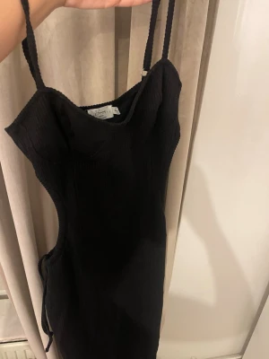 Svart ribbad kort klänning med snörning - Svart ribbad klänning med smala axelband och snörning i sidorna. Snygg, figurnära modell med croppad känsla och stretchigt material. Perfekt för dig som gillar minimalistisk stil med en twist.