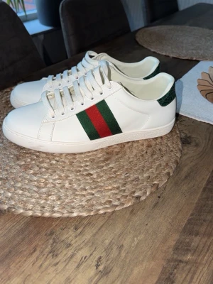 Gucci vita sneakers med gröna/röda ränder - Snygga vita sneakers från Gucci i läder med klassiska gröna och röda ränder på sidan samt grön detalj vid hälen. Skorna har vita snören och en stilren, låg profil som passar till det mesta. Perfekta för dig som gillar exklusiv och trendig streetstyle. Medföljer endast skorna!