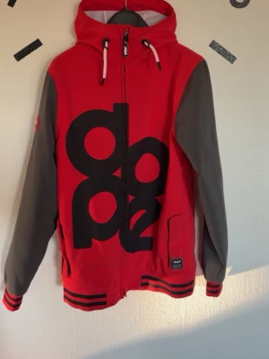 Röd vindjacka med huva från Dope - Röd vindjacka från Dope med stora svarta tryck på framsidan, grå ärmar och huva med dragsko. Jackan har hel dragkedja, ribbade muddar med svarta ränder och fickor på sidorna. Perfekt för vår och höst.