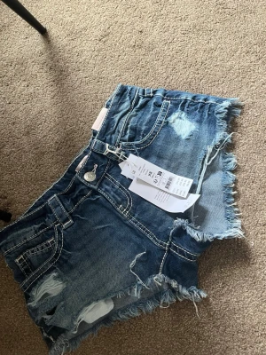 Blå jeansshorts med fransig kant - Säljer ett par blå jeansshorts från Gina tricot med fransig kant och slitna detaljer. Shortsen har klassisk femficksmodell, synliga sömmar och råa kanter för en cool look. Perfekta för varma dagar och enkel att matcha med allt.