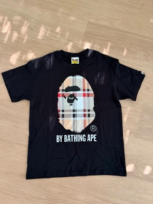 Svart BAPE t-shirt med rutigt tryck - Svart t-shirt från BAPE med det ikoniska apansikte-trycket i rutigt mönster på bröstet och texten 'BY BATHING APE' under. Klassisk passform och rund halsringning. Perfekt för dig som gillar streetwear och vill sticka ut.