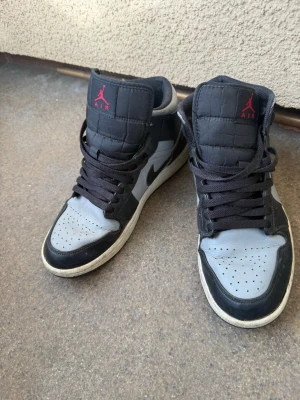 Nike Air Jordan 1 Mid svart/grå - Säljer ett par Nike Air Jordan 1 Mid i svart och grått med klassisk röd Jumpman-logga på plösen. Skorna har svart swoosh, svarta skosnören och perforerad tåbox. Tillverkade i läder och textil för en ikonisk streetstil.