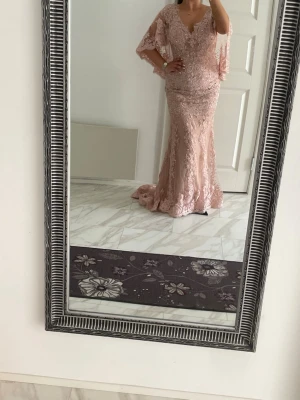 Lång rosa spetsklänning med volangärm balklänning  - Elegant långklänning i ljusrosa spets med v-ringning och volangärmar. Klänningen har figurnära passform och ett vackert fallande släp. Perfekt för bal eller festliga tillfällen. Detaljerad spets och romantisk look.