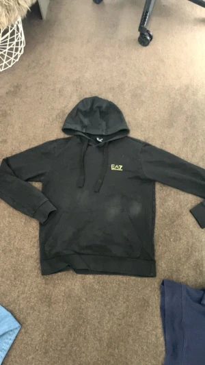 Svart hoodie från EA7 Emporio Armani - Säljer en svart hoodie från EA7 Emporio Armani med gul logga på bröstet och gul detalj på ryggen. Klassisk modell med huva och dragsnören, samt ribbade muddar vid ärmslut och nederkant. Perfekt för en avslappnad och stilren look.