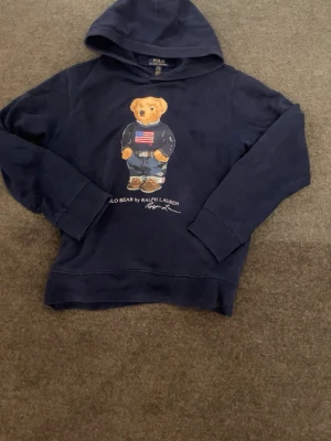 Marinblå hoodie Polo Bear Ralph Lauren - Snygg marinblå hoodie från Ralph Lauren med den ikoniska Polo Bear-trycket på bröstet. Hoodien har huva, långa ärmar och ribbade muddar. Materialet är mjukt och bekvämt, perfekt för en avslappnad stil. Klassisk design med en cool björn i jeans och flaggtröja.