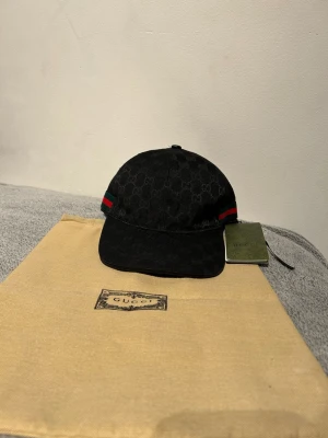 Svart keps från Gucci med GG-mönster - Snygg svart keps från Gucci med klassiskt GG-monogram över hela kepsen. Har de ikoniska gröna och röda ränderna på sidan och justerbart spänne bak. Tillverkad i canvasmaterial och levereras med originalpåse och tagg.