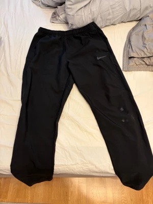 Svarta woven pants från Nike - Säljer ett par svarta woven pants från Nike med avslappnad passform och resår i midjan. Byxorna har en diskret Nike-logga på vänster lår och är tillverkade i ett mjukt och bekvämt material, perfekt för chill dagar eller träning.