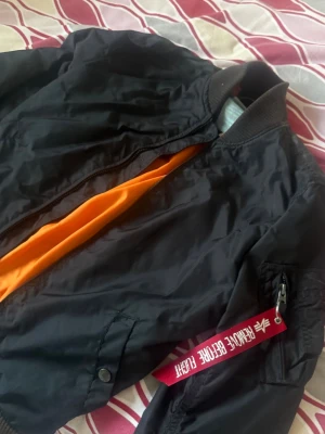 Svart bomberjacka Alpha Industries - Klassisk svart bomberjacka från Alpha Industries med orange innerfoder och ribbade muddar. Jackan har dragkedja, ficka på ärmen med röd tagg 'Remove Before Flight' och tryckknappar på fickorna. Perfekt för en streetwear-look. Kan mötas upp i Helsingborg och Malmö , vid leverans tillkommer pris kan sänka varans pris vid snabb äffär 