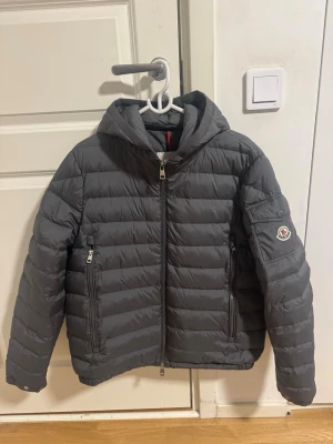 Grå dunjacka med huva från Moncler - Snygg grå dunjacka från Moncler med huva och dragkedja framtill. Jackan har två framfickor med dragkedja och Moncler-logga på ärmen. Perfekt för kalla vinterdagar och ger en stilren look. Klassisk quiltad design och hög krage för extra värme.