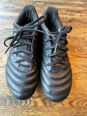 Svarta fotbollsskor Copa från Adidas - Säljer ett par svarta fotbollsskor från Adidas med klassisk design och snörning. Skorna har en mönstrad ovandel i syntetmaterial och textil, samt diskreta Adidas-ränder på sidan. Perfekta för dig som vill ha stilrena och funktionella skor på planen.