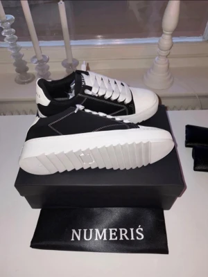Numeris Skor - Snygga sneakers från Numeris med svart ovandel och vita detaljer. Skorna har kraftig vit sula med räfflad design, vita skosnören och kontrasterande sömmar. Perfekt för dig som vill ha en modern och stilren look.