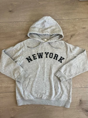 Grå hoodie med New York-tryck - Säljer en grå hoodie med svart New York-tryck på bröstet, den är från H&M och ör i storleken xs. Hoodien har huva med dragsko, ribbade muddar och en stor magficka. Perfekt för dig som gillar streetstyle och vill ha något bekvämt och snyggt. Materialet är mjukt och känns skönt mot huden.