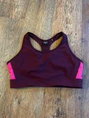 Vinröd sport-bh med rosa detaljer - Snygg vinröd sport-bh från energetics med kontrasterande rosa paneler på sidorna. Bh:n har brottarrygg och bred resår under bysten för extra stöd. Tillverkad i ett stretchigt och snabbtorkande material, perfekt för träning och sport. Använd 2 gånger