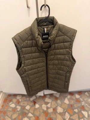 Olivgrön dunväst från BOSS - Snygg olivgrön dunväst från BOSS med quiltad design och hög krage. Västen har dragkedja framtill och diskret logotyp på bröstet. Perfekt för lager-på-lager och håller dig varm under kyliga dagar. Materialet är lätt och smidigt, vilket gör den enkel att bära.