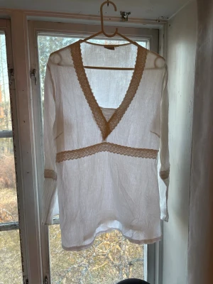 Vit blus - Säljer en vit blus med djup V-ringning och spetsdetaljer längs halsen, midjan och ärmsluten. Blusen är tunn och har en luftig känsla, perfekt för lager-på-lager. Långärmad och tillverkad i ett lätt material som ger en avslappnad look.