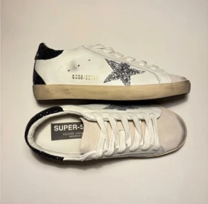 Golden Goose sneakers - Golden Goose Super-Star sneakers i vitt läder med silverglittrig stjärna på sidan och svart glitterdetalj på hälen. Beige sula med vintage-look och vita skosnören. Klassisk låg modell med rund tå och perforerade detaljer. Jättebra skick, pris kan diskuteras vid snabb affär 