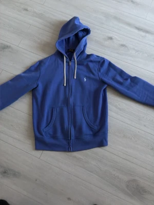 Blå hoodie från Polo Ralph Lauren - Säljer en blå hoodie från Polo Ralph Lauren med vit logga på bröstet och vita snören i huvan. Hoodien har dragkedja, två fickor fram och är tillverkad i mjukt bomullsmaterial. Perfekt för dig som vill ha en klassisk och stilren look.