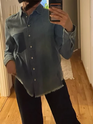 jeansskjorta  - Säljer en ljusblå jeansskjorta i klassisk modell med knappstängning framtill och en stor bröstficka (ej riktig ficka). 