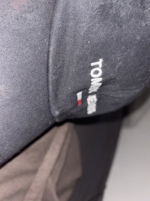 Svart keps från Tommy Jeans - Snygg svart keps från Tommy Jeans med broderad logga och den klassiska lilla flaggan framtill. Justerbart spänne bak och gröna detaljer på insidan. Perfekt accessoar för en casual och trendig look.